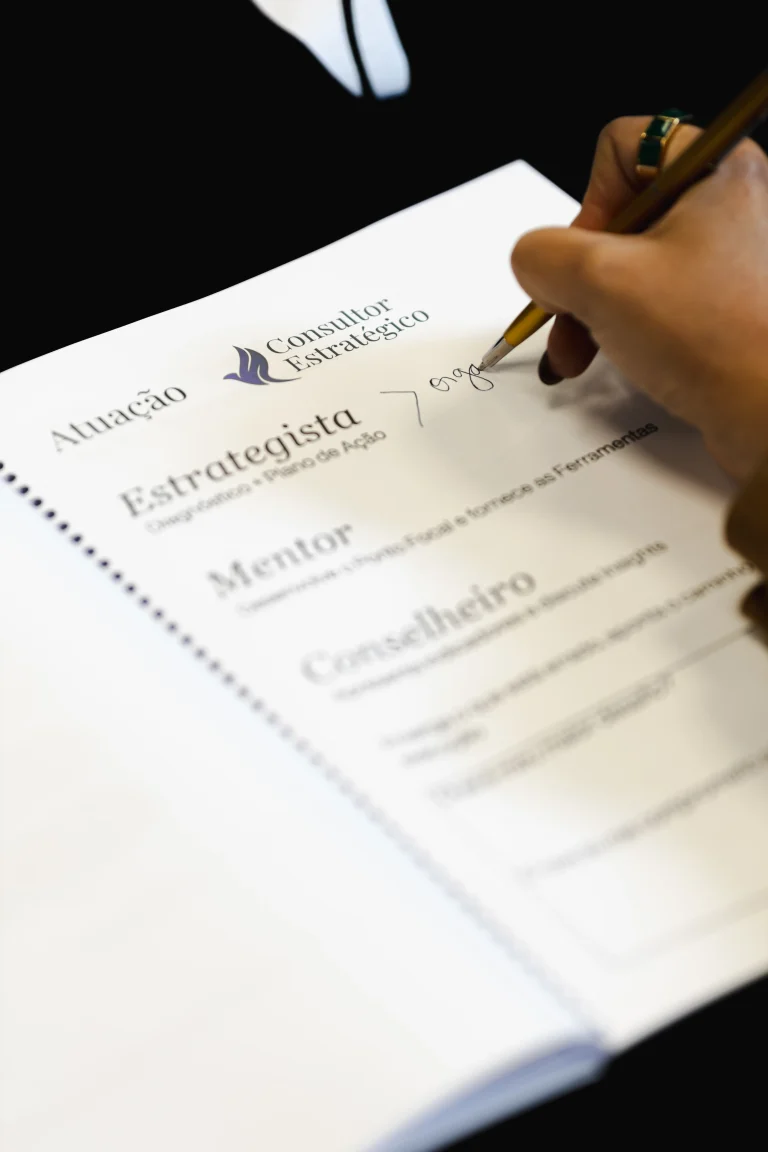 Mão feminina com anel dourado escrevendo a lápis sobre a apostila 'Consultor Estratégico' aberta — logo e pássaro símbolo visíveis, categorias 'Estrategista, Mentor, Conselheiro' impressas na página